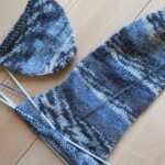 Halbfertige blau melierte Socke auf einem Nadelspiel neben einer fertigen Socke