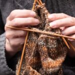 Hände stricken eine Socke aus brauner Wolle mit einem Nadelspiel